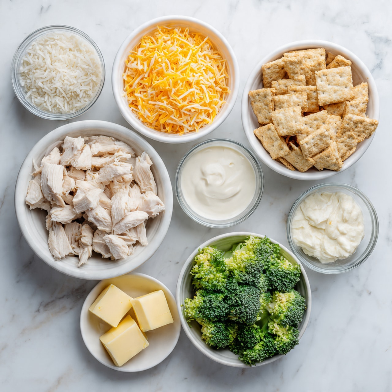 This image shows seven bowls on a white marbled surface. Starting from the top left, there is a small bowl with a white creamy sauce. To the right, a white bowl filled with broken light brown crackers. Below the crackers, a small white bowl contains three cubes of yellow butter. In the middle right, a white bowl holds chunks of cooked chicken with a light beige color. On the bottom right, a white bowl is filled with fresh green broccoli florets. In the bottom left corner, a small bowl contains a thick white sauce. Above it, a white bowl is filled with shredded orange cheddar cheese. In the center left, a clear glass bowl holds cooked white rice. Photo taken with an iphone --ar 4:5 --v 7
