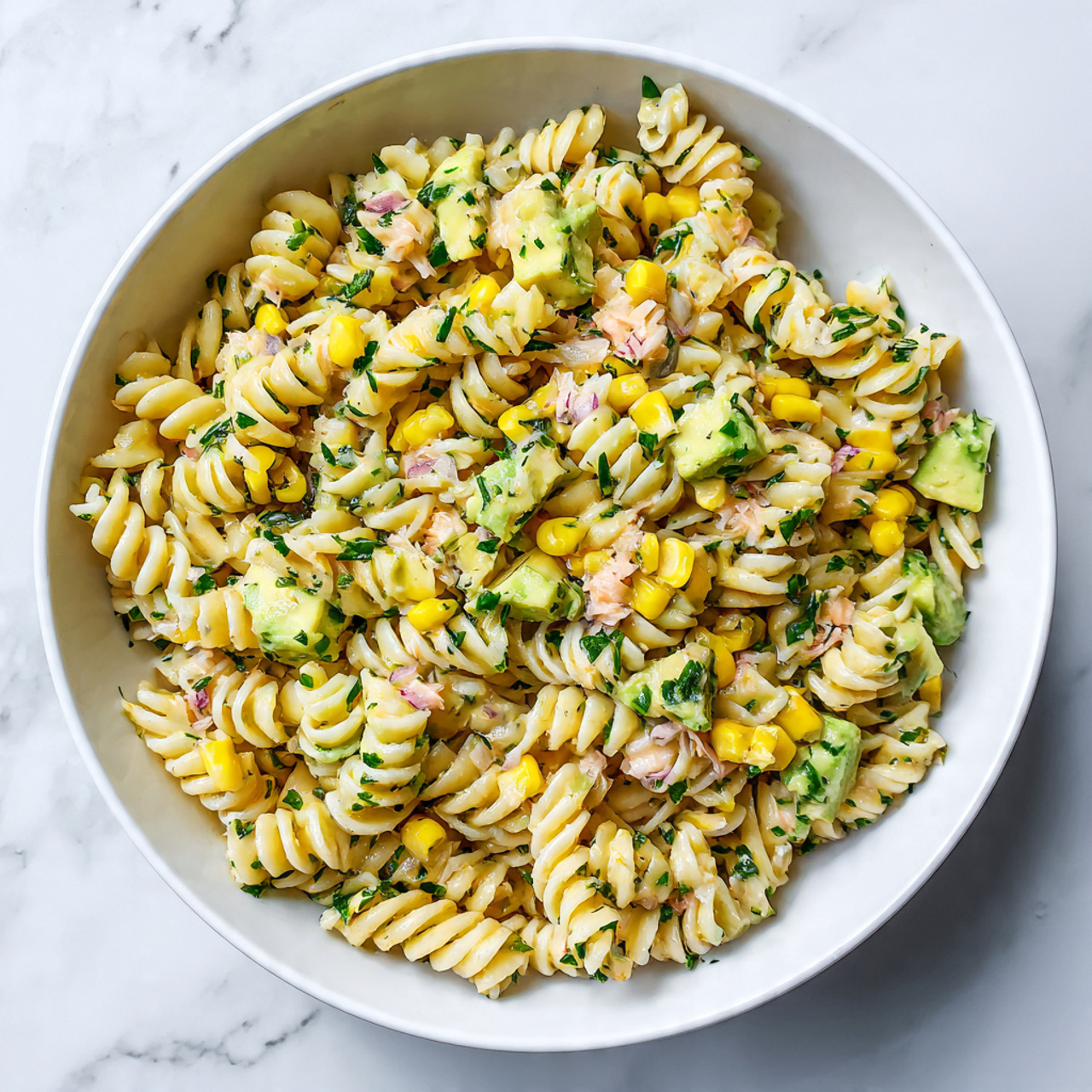 The image shows a white bowl filled with a pasta salad made of spiral pasta mixed with yellow corn kernels and small green avocado cubes scattered evenly throughout. There are small pink pieces, likely crab or seafood, adding touches of color mixed with finely chopped green herbs. The pasta looks firm but soft, while the corn and avocado add a fresh texture. The white bowl sits on a white marbled surface. Photo taken with an iphone --ar 4:5 --v 7