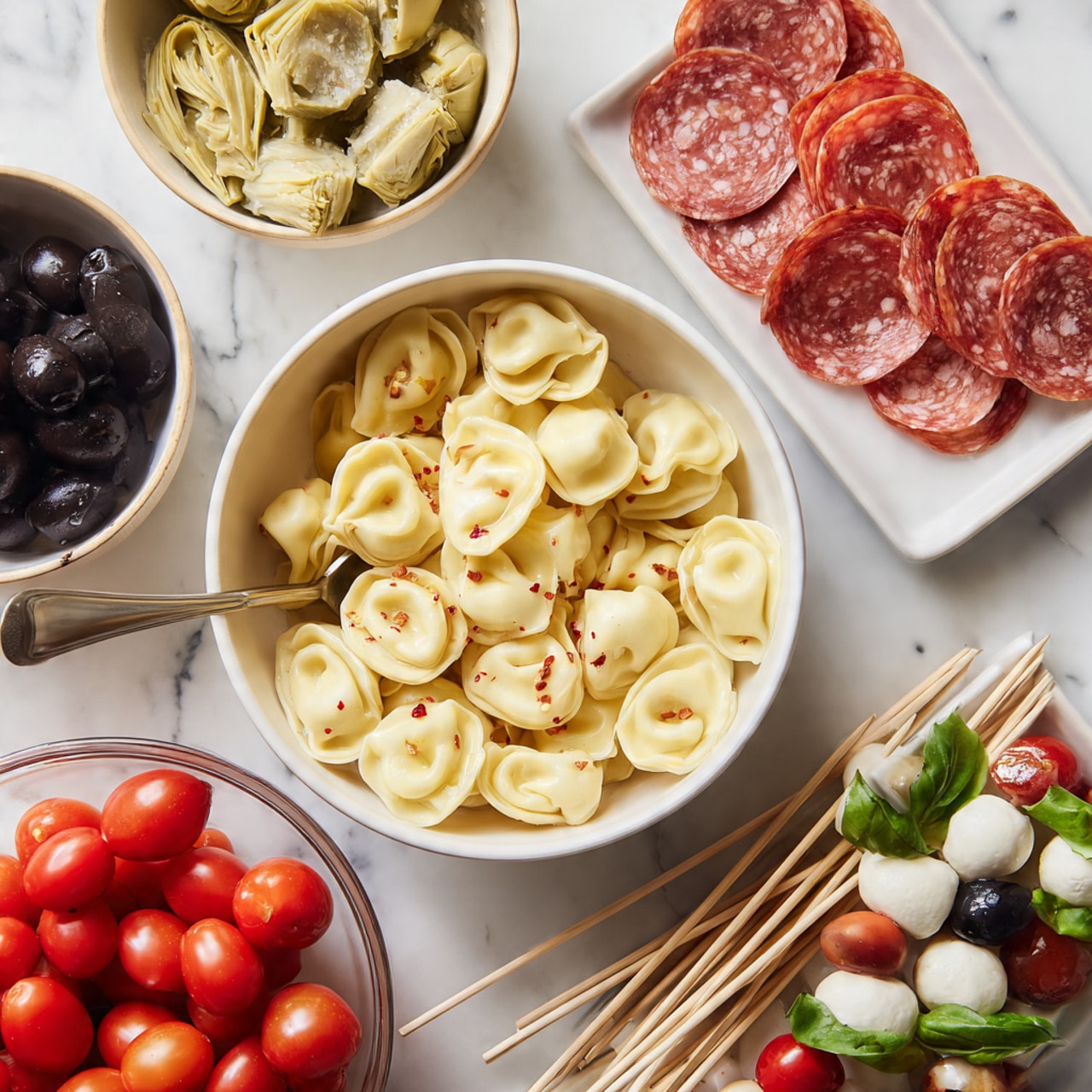 A white bowl filled with pale yellow tortellini pasta with red pepper flakes, a silver spoon resting inside the bowl; above it is a beige bowl with light green artichoke hearts in oil; to the right are scattered pink and white slices of salami next to a white bowl of shiny black olives; a white rectangular plate near the top right holds skewers made of red cherry tomatoes, white mozzarella balls, green basil leaves, light beige mushrooms, and dark olives; at the bottom left, a glass bowl filled with red cherry tomatoes and a bunch of wooden skewers lie next to a pair of woman's hands threading a slice of salami onto a skewer that already holds a mushroom, mozzarella ball, basil leaf, and tomato; all items rest on a white marbled surface photo taken with an iphone --ar 4:5 --v 7
