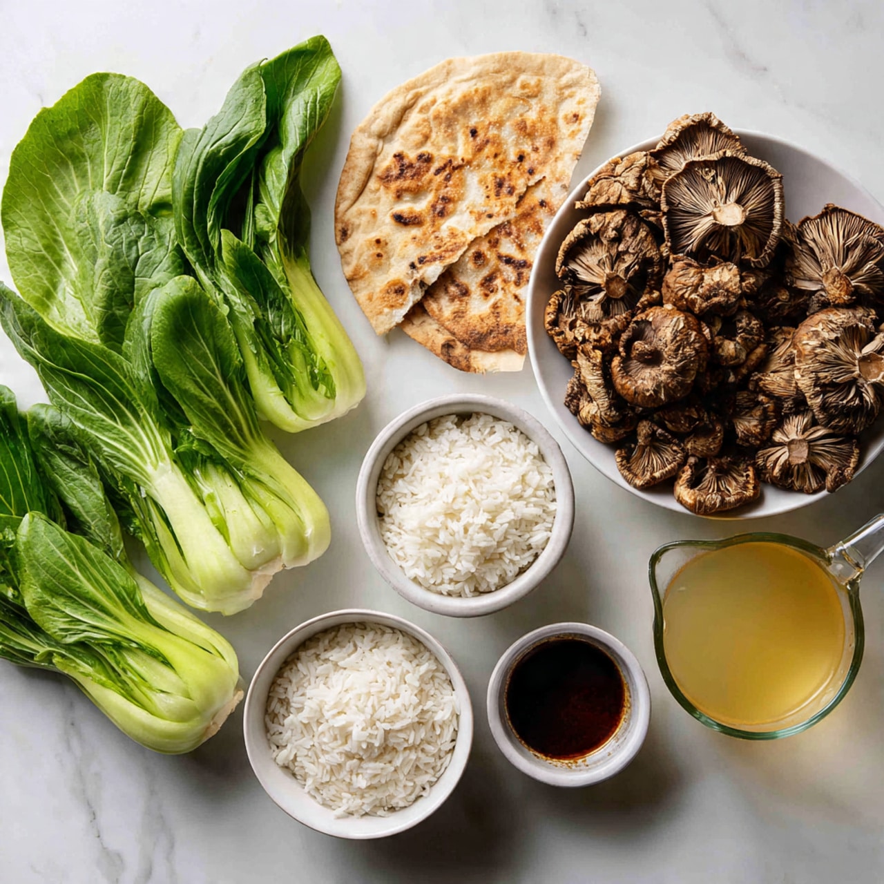 The image shows a collection of fresh ingredients placed on a white marbled surface. At the back left, there are fresh, bright green bok choy leaves with crisp white stalks. Next to them, on the right, is a white bowl filled with large, brown shiitake mushrooms showing their textured caps and gills. Above the mushrooms, there is a piece of flatbread with a light brown color. Below the bok choy, two white bowls hold white rice grains, one larger bowl and one smaller bowl. In between the rice bowls and the mushrooms, there is a small white dish filled with dark brown sauce. To the right of all these, there is a clear glass measuring cup filled with a pale yellow broth. photo taken with an iphone --ar 4:5 --v 7