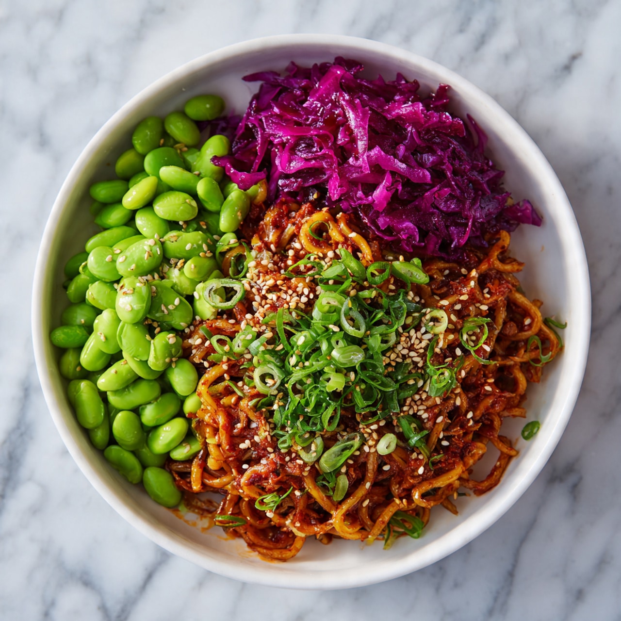 The image shows a white bowl filled with three main sections of food. The front half is full of cooked curly noodles, coated in a reddish sauce and sprinkled with sesame seeds and chopped green onions. On the left side of the bowl, there is a neat pile of bright green edamame beans. The top right corner has a small portion of vibrant purple pickled cabbage. The bowl is placed on a surface with a white marbled texture. Photo taken with an iphone --ar 4:5 --v 7