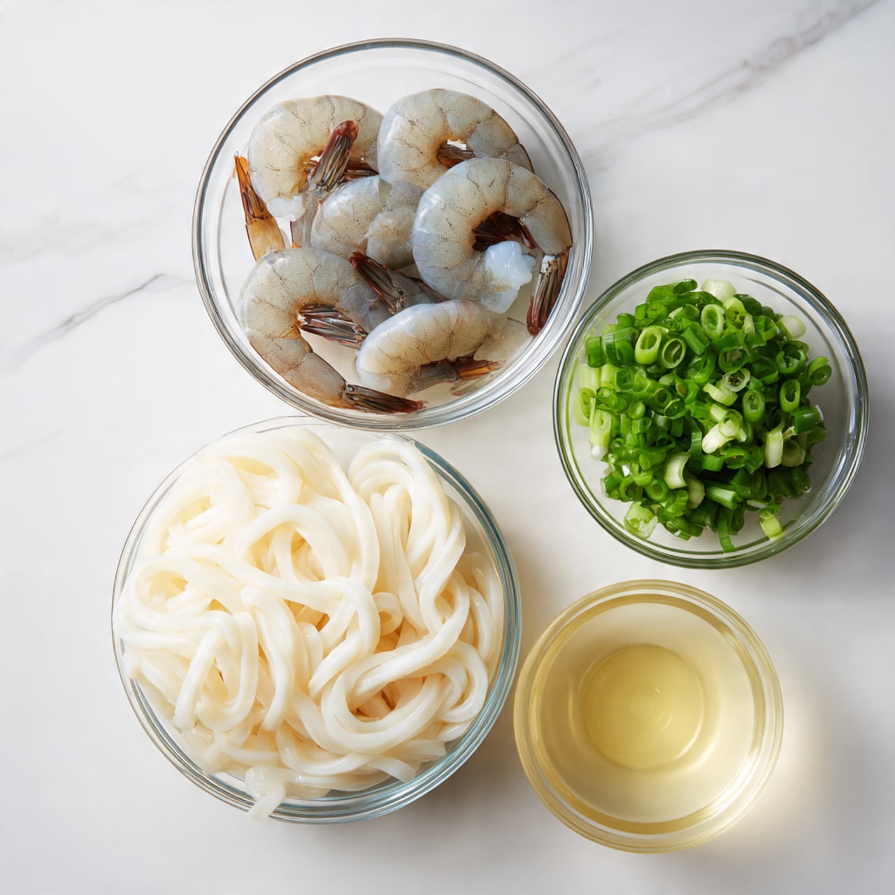 The image shows four clear glass bowls on a white marbled surface. The largest bowl in the front holds a single block of thick, white udon noodles with a smooth, slightly glossy texture. To the left, a medium bowl contains five raw shrimp that are pale grey with darker brown tails, resting in a circle. Behind them, a smaller bowl holds chopped green onions, bright green and white in color with a fresh look. The smallest bowl on the right contains a pale yellow liquid, likely cooking oil or broth. The bowls are arranged close together in a neat, simple layout. Photo taken with an iphone --ar 4:5 --v 7