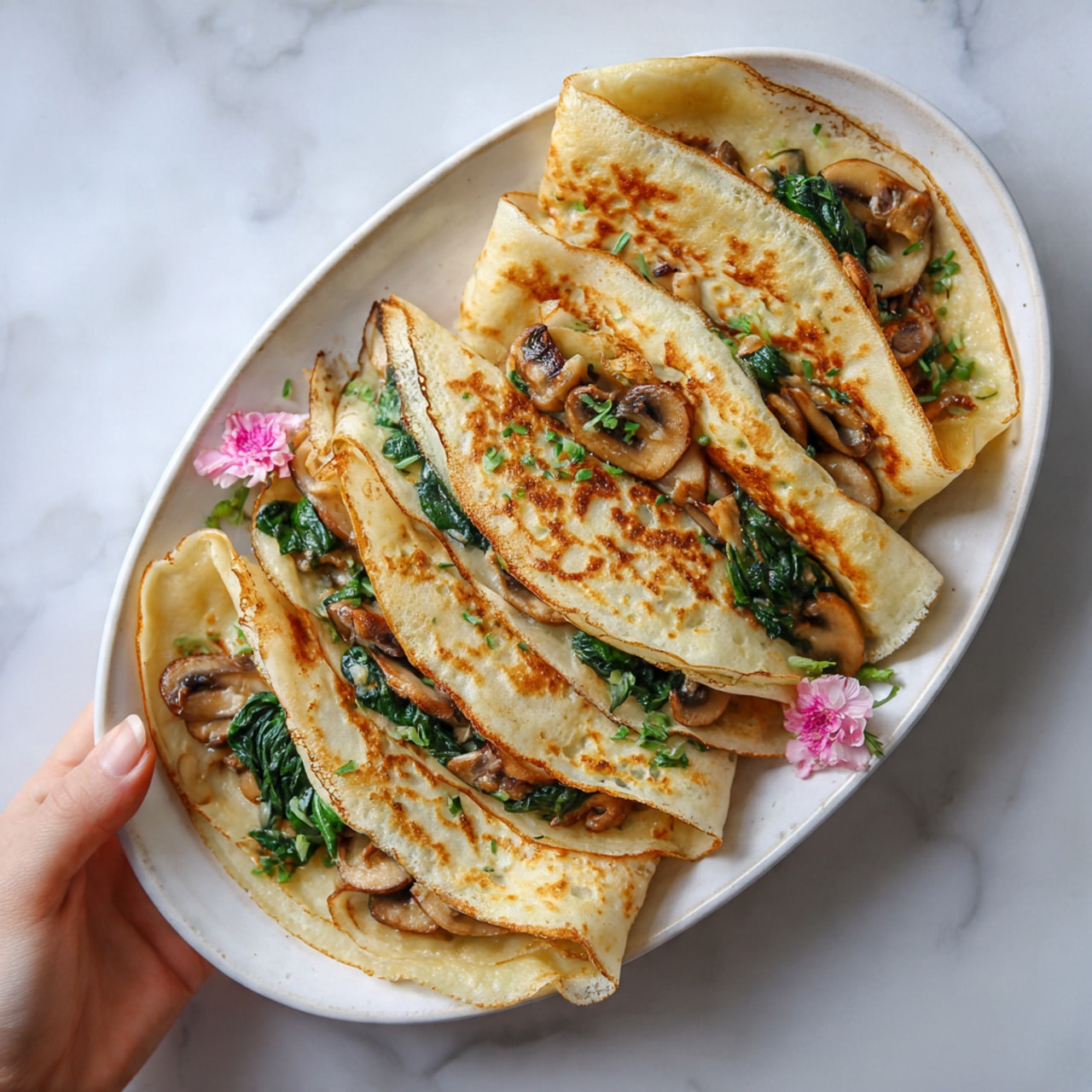 The image shows a white oval plate on a white marbled surface, filled with several folded crepes stacked slightly on top of each other. Each crepe is light golden brown with some crispy darker spots, and inside the crepes are visible layers of sautéed spinach and slices of light brown mushrooms. Two small pink flowers lie on the bottom left side of the plate, adding a gentle decoration. A woman's hand is holding one crepe from the left side, showing the texture of the soft crepe and the filling inside. Photo taken with an iphone --ar 4:5 --v 7