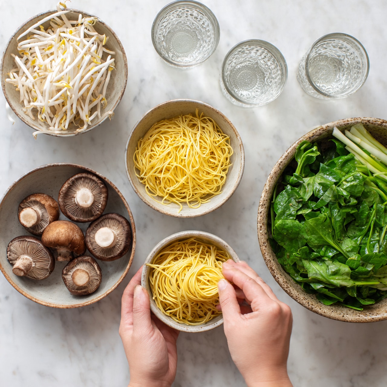 The image shows five white bowls on a white marbled surface. The front left bowl is filled with yellow cooked noodles, with a woman's hand holding some noodles above it. To its front right are three brown mushrooms with visible rings and a white base. Behind them on the right is a large bowl full of vibrant green leafy vegetables and whole scallions. To the back left is a bowl of bean sprouts appearing white and fresh. Next to it are three empty glass cups. The scene looks natural and ready for cooking photo taken with an iphone --ar 4:5 --v 7