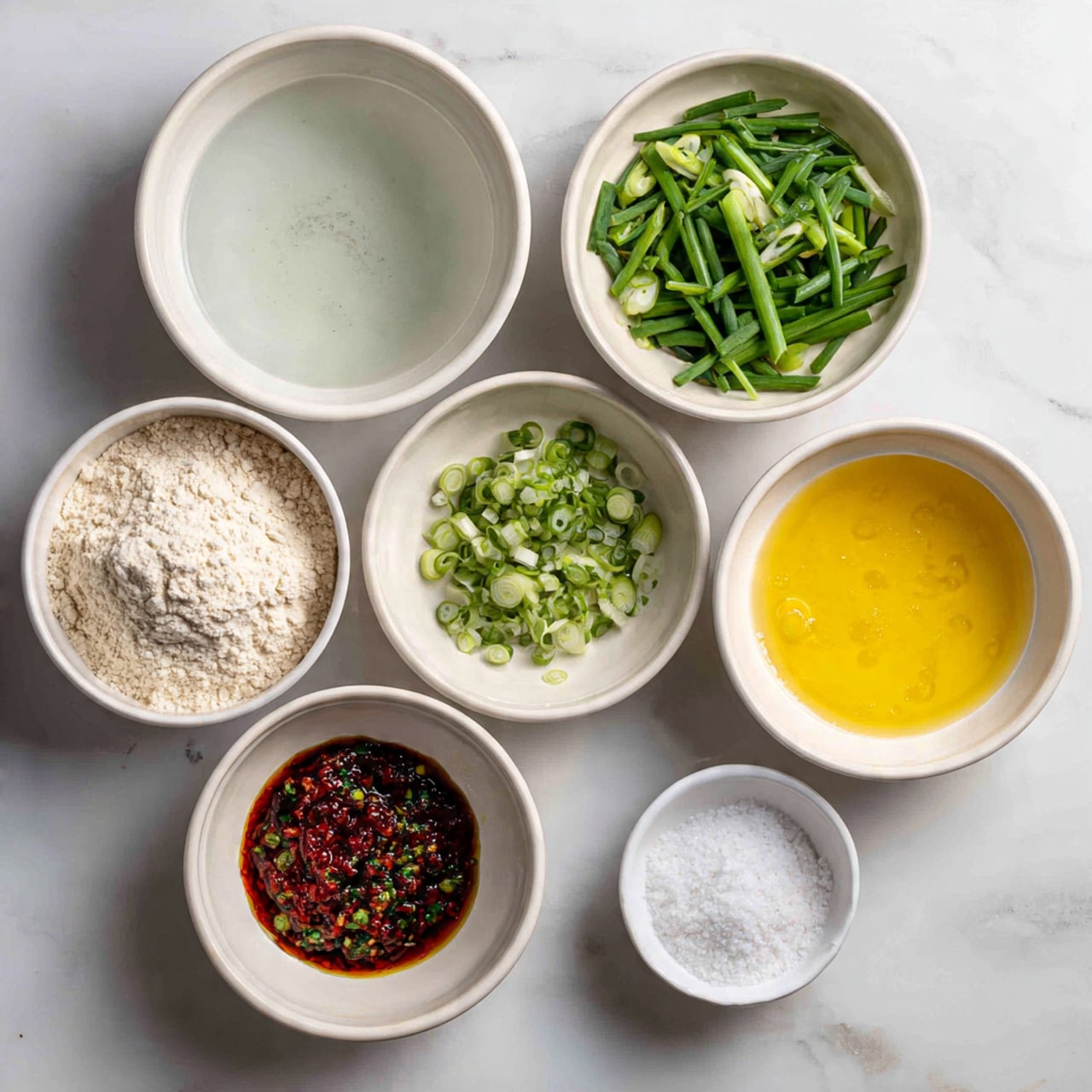 The image shows seven white bowls of different sizes arranged on a white marbled surface. The largest bowl on the left contains water with a clear, smooth texture. Below it is a bowl filled with light beige flour, which looks powdery with a slightly clumpy texture. At the top middle is a bowl with chopped green onions, showing bright green stalks cut into pieces. To the right of the green onions is a bowl of golden yellow oil, smooth and shiny. Below that is a bowl with a dark red and green spicy sauce that looks oily and textured with chopped ingredients. Under this bowl, there is a bowl with a yellow beaten egg mixture that looks slightly frothy. Finally, on the far right bottom is a small white bowl with white salt crystals. Photo taken with an iphone --ar 4:5 --v 7