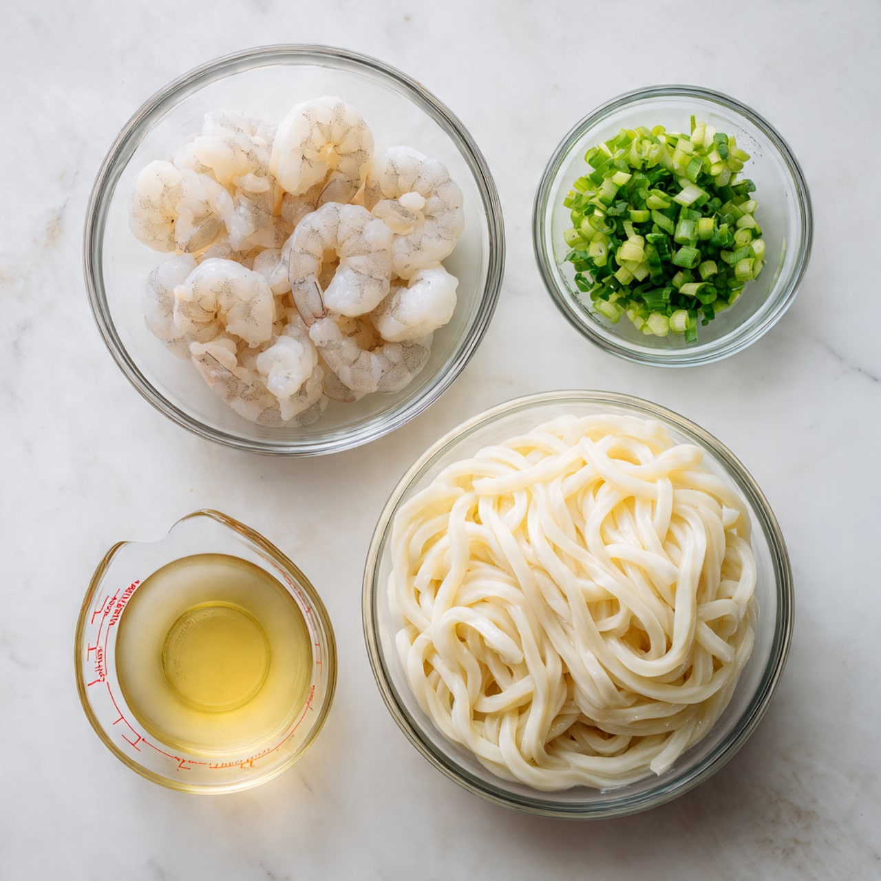The image shows four clear glass bowls on a white marbled surface. The largest bowl at the bottom right contains a nest of thick, white udon noodles with a soft texture tightly packed in a block. Above this, to the left, a medium-sized bowl holds several raw, peeled shrimp with a pale pink and white color, arranged loosely. To the top right of the shrimp, a small bowl contains chopped green onions, showing bright green and white pieces. Finally, to the far right, a small measuring cup holds a light yellow liquid, likely oil or broth, with red measurement markings visible on its surface. photo taken with an iphone --ar 4:5 --v 7