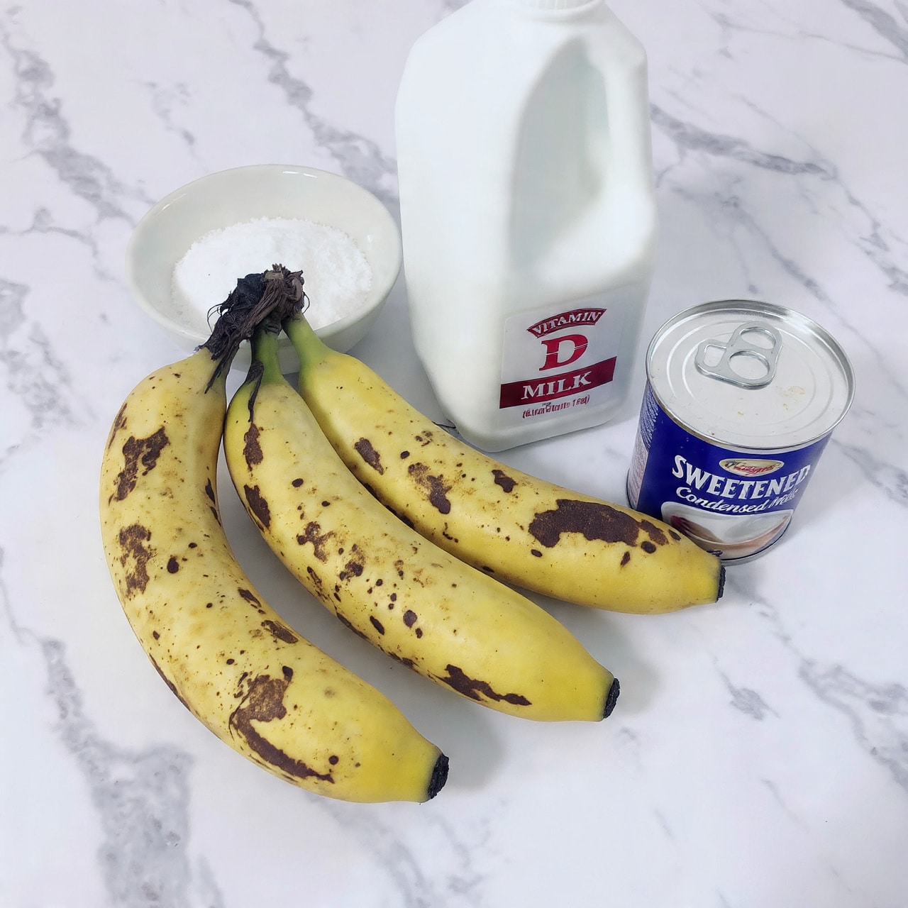 The image shows four ripe bananas with many brown spots on their yellow skins, placed on a white marbled surface. Next to the bananas, there is a white bowl full of white granulated sugar. Behind the bowl, there is a white plastic jug of milk with a red and white label that reads