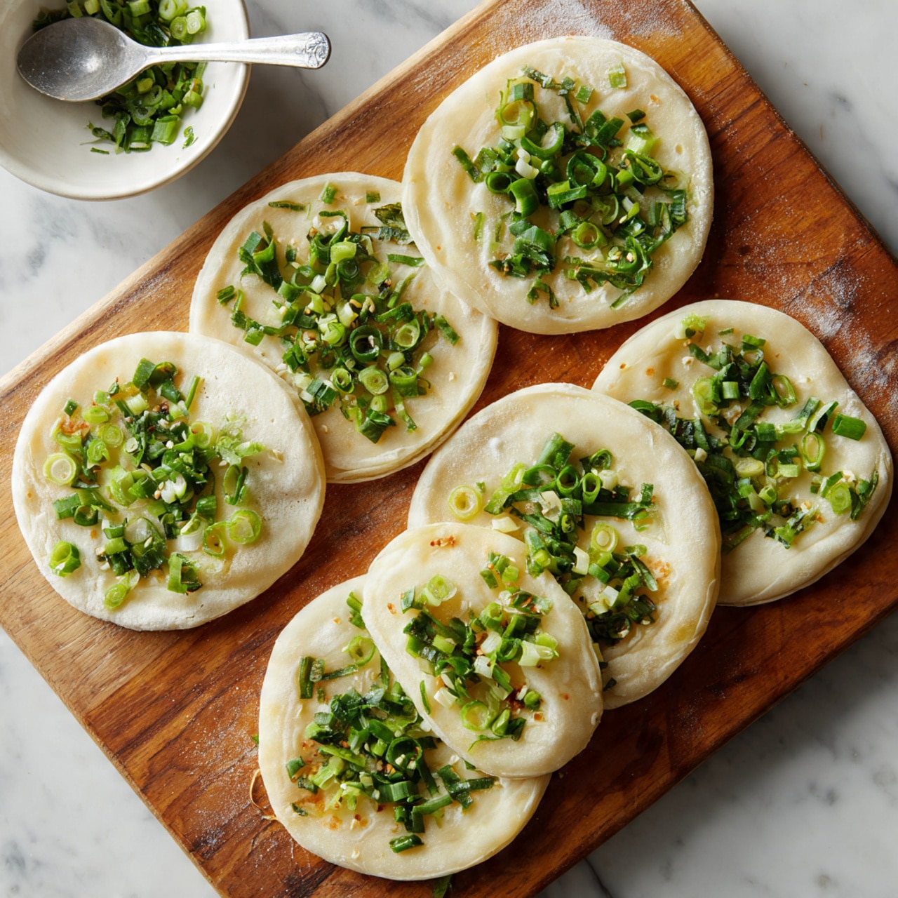 Six circular pieces of thin dough lie flat on a wooden board, each topped with chopped green onions spread mostly in the middle. The dough circles are light beige and smooth, slightly shiny from moisture. The green onions add a fresh bright green color, scattered in small chunks on each piece. There is a white bowl with a metal spoon blurred at the corner. The scene is set on a white marbled surface. photo taken with an iphone --ar 4:5 --v 7
