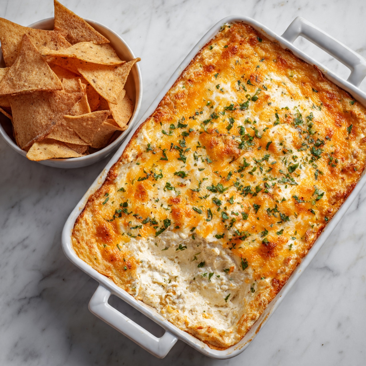 The image shows a white ceramic rectangle dish filled with a baked cheesy dip. The top layer is golden orange melted cheese sprinkled with finely chopped green herbs. One corner of the dip is scooped out, revealing a creamy, slightly chunky white inside. Next to the dish, there is a white bowl full of triangular toasted chips. The dish and bowl sit on a white marbled surface. photo taken with an iphone --ar 4:5 --v 7