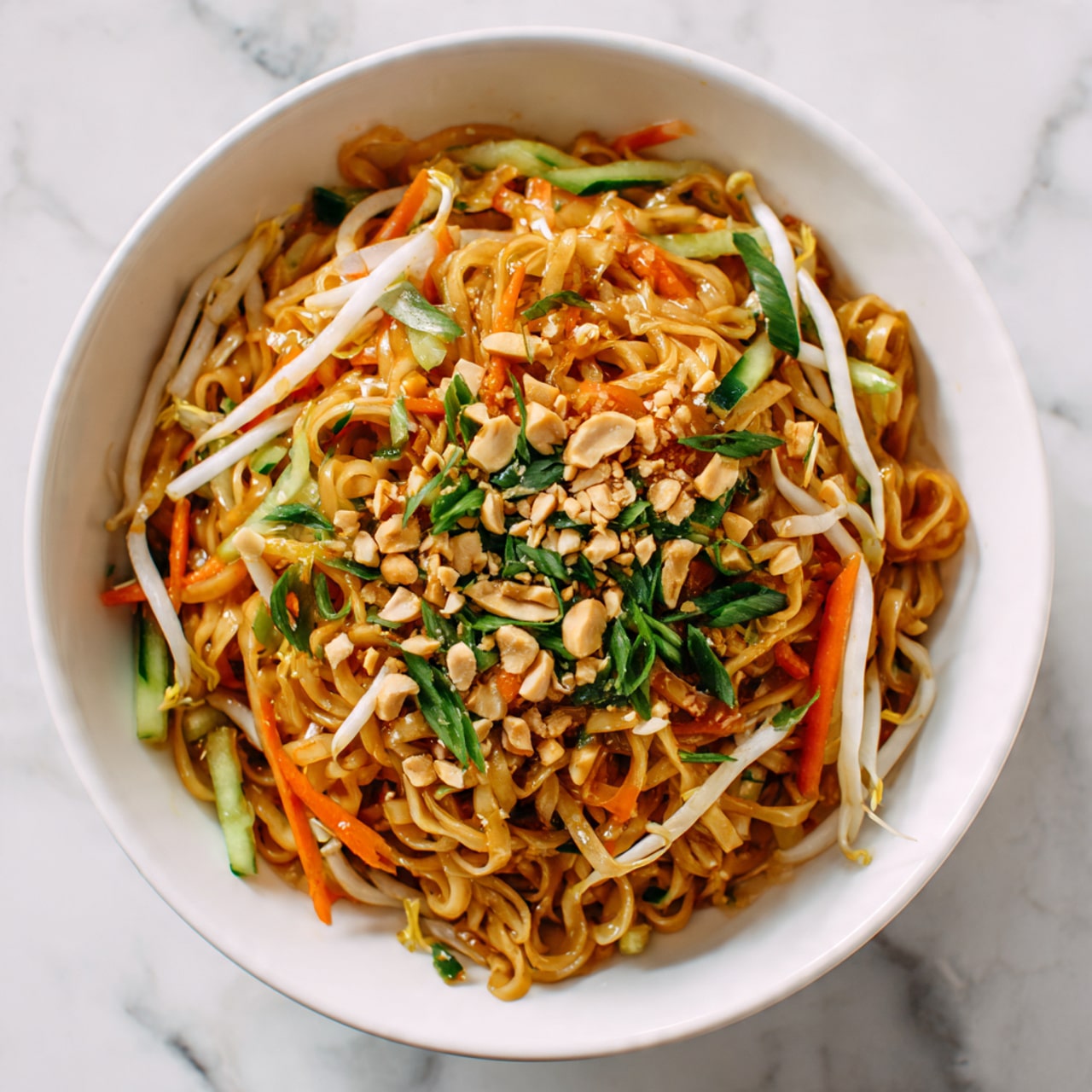 The image shows a white bowl filled with a noodle dish. The bottom layer is light brown cooked noodles mixed with a glossy sauce, giving them a slightly shiny texture. Scattered throughout the noodles are thin strips of orange carrot, white bean sprouts, and green cucumber, all fresh and crisp looking. Small chopped green herbs are sprinkled on top, adding a touch of brightness. Some crushed light brown peanuts are scattered unevenly over the dish. The bowl sits on a white marbled surface. photo taken with an iphone --ar 4:5 --v 7