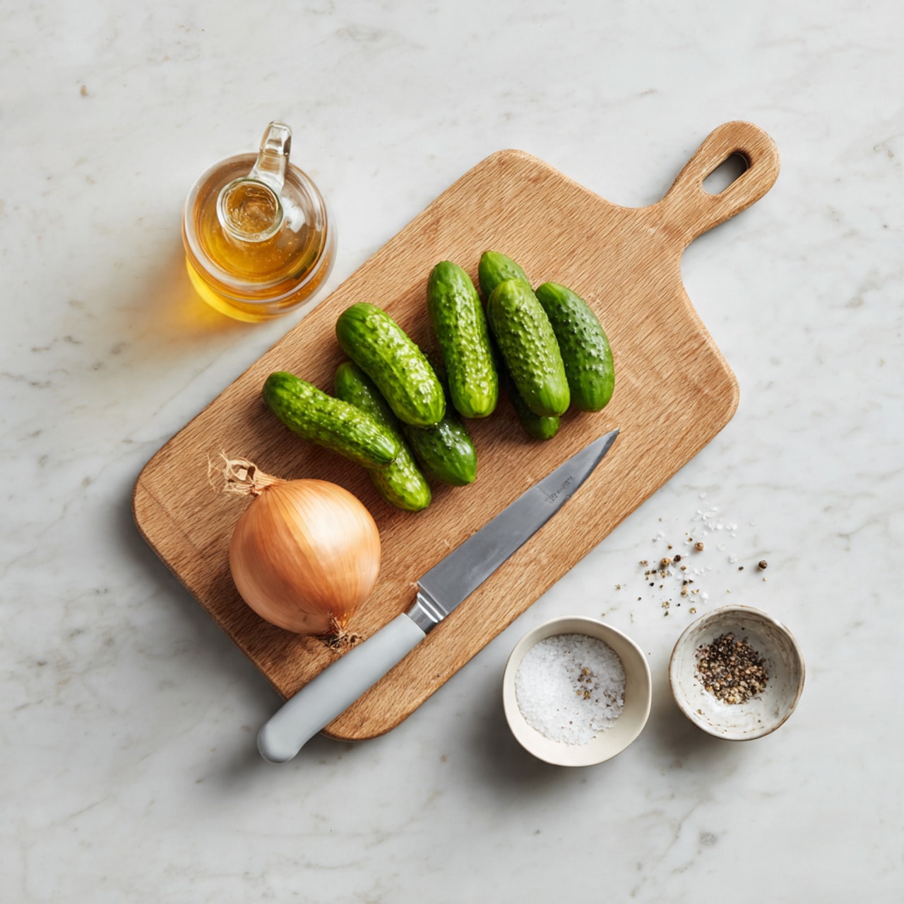 The image shows a wooden cutting board placed on a white marbled surface. On the cutting board, there is a light brown shallot, several bright green small cucumbers, a knife with a light grey handle, a small white bowl filled with salt, and a small white bowl with black pepper. Next to the cutting board, there is a clear bottle with a golden liquid inside. The arrangement is neat and well-organized. Photo taken with an iphone --ar 4:5 --v 7