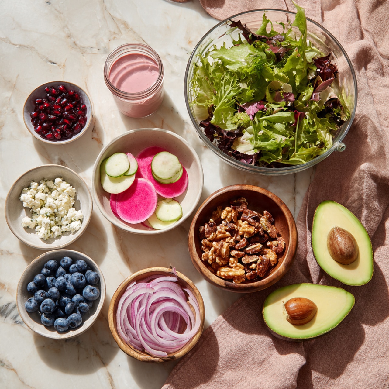 The image shows a mix of fresh ingredients arranged on a white marbled surface. At the top right, there is a large clear glass bowl filled with mixed green leafy salad. To the left of it, a small clear jar contains a pink dressing. Below, there are seven smaller white bowls and one wooden bowl. The white bowls hold fresh blueberries, pink and green round slices of radishes, deep red pomegranate seeds, thin light purple onion slices, and crumbled white cheese. The wooden bowl contains roasted nuts that are golden brown and slightly shiny. To the right on the marbled surface, there are two avocado halves, one with the seed and one without, sitting on a dusty pink cloth. photo taken with an iphone --ar 4:5 --v 7