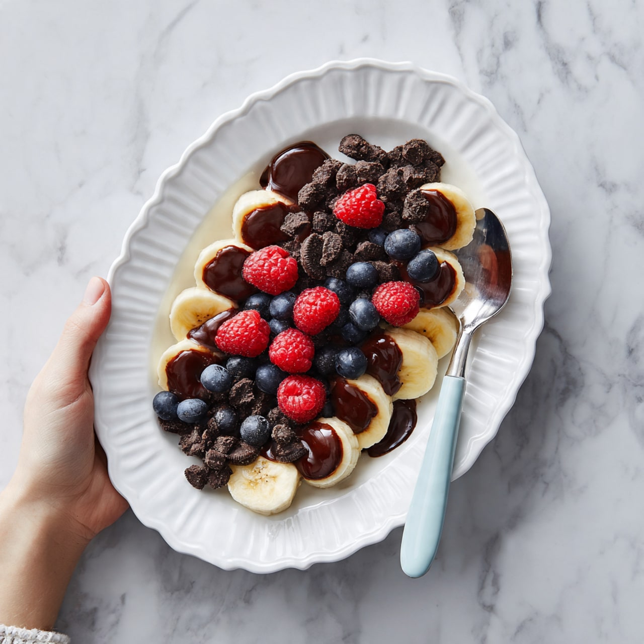 The image shows a white scalloped oval plate filled with a dessert made of three layers. The bottom layer consists of halved bananas placed side by side along the plate’s length. On top of the bananas, there is a layer of small pieces of dark chocolate cookies, unevenly spread. The next layer is colorful fresh berries, including red raspberries and dark blueberries scattered across. The top layer is a thick dark chocolate sauce drizzled over the berries and cookies. A white spoon with a light blue handle is placed on the right side of the plate on a white marbled surface. A woman's hand holding the plate is visible at the upper left corner. Photo taken with an iphone --ar 4:5 --v 7