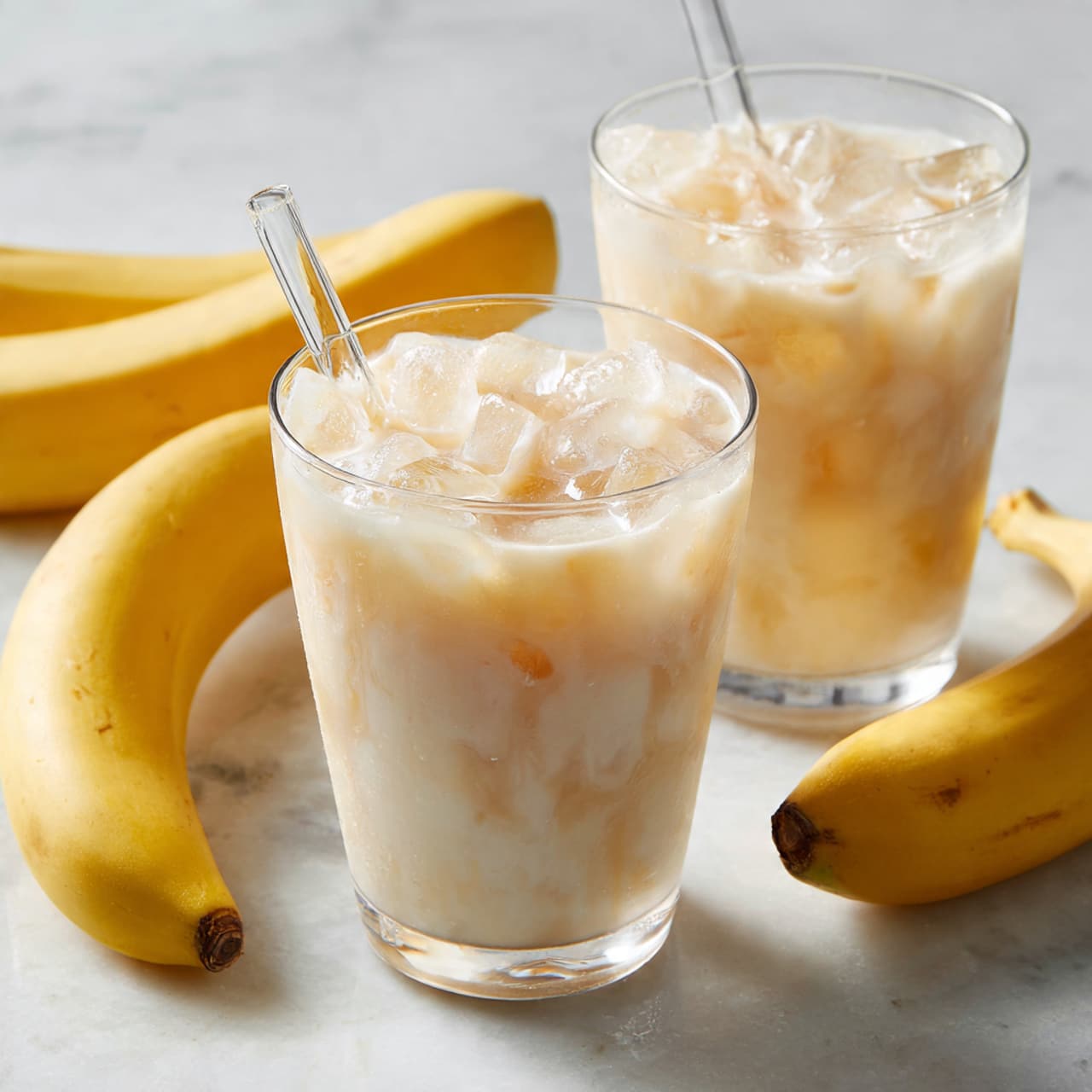 Two clear glass cups filled with a creamy light beige drink with ice cubes that create soft white and translucent layers on top. One cup is in front-center, showing a clear glass straw inside, while the other is placed slightly behind and to the right. Around the glasses, there are several whole yellow bananas with a smooth texture, one partially peeled laying flat. The entire setup is on a white marbled surface with a soft, blurred background. photo taken with an iphone --ar 4:5 --v 7