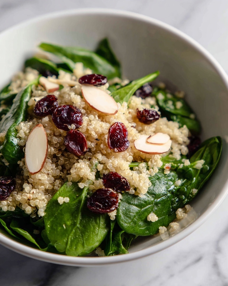 The image shows a close-up of a fresh quinoa salad in a white bowl. The salad has several layers, starting with a base of fluffy, off-white quinoa grains spread evenly. On top, there are bright green spinach leaves mixed throughout, adding a fresh, leafy texture. Dark red dried cranberries are scattered across the surface, providing contrast and small pops of color. Thin slices of almond, pale beige in color, are placed on different spots, adding a smooth, layered look. The salad is vibrant, fresh, and colorful, set against a white marbled surface. photo taken with an iphone --ar 4:5 --v 7