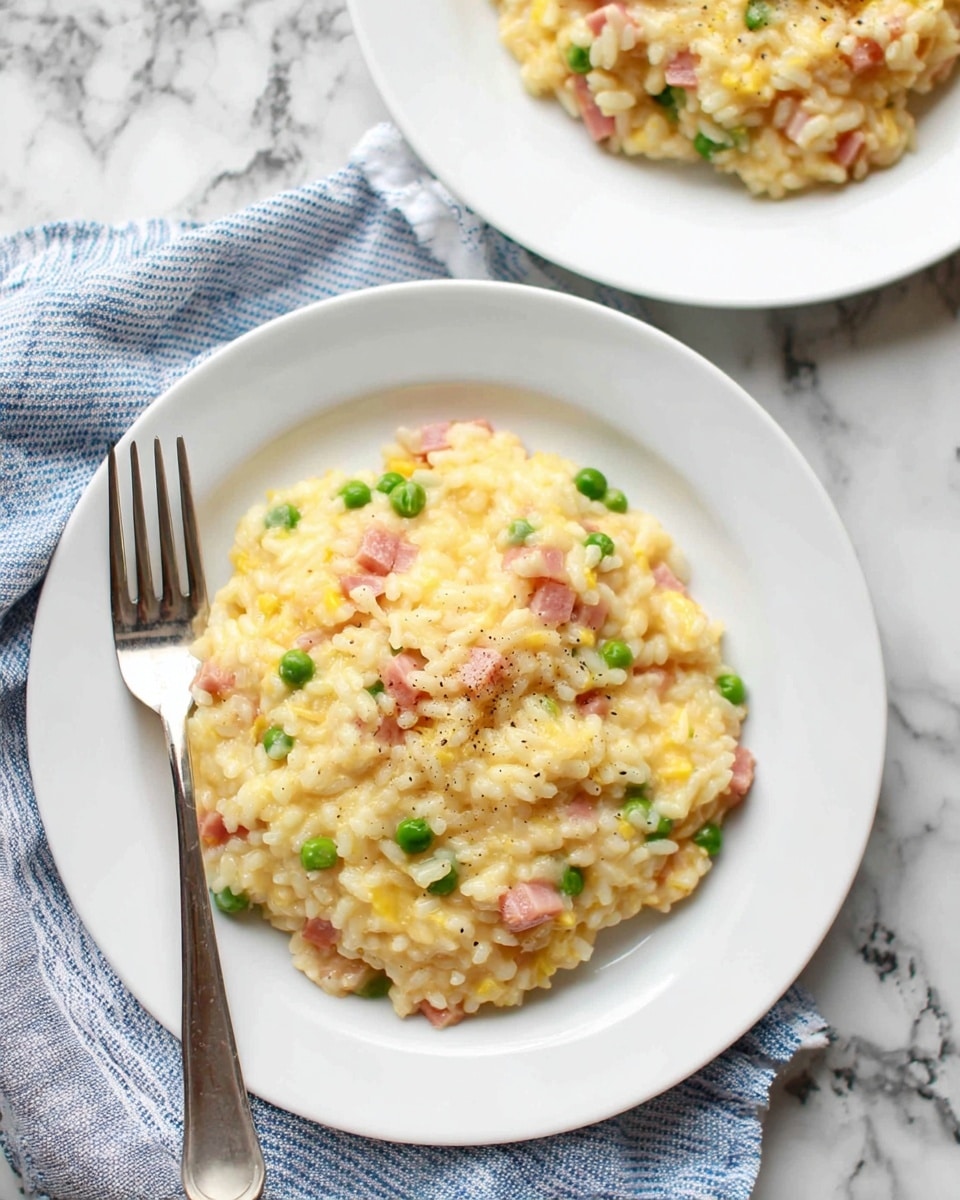 The image shows a white plate with a creamy risotto dish served on a white marbled surface. The risotto has a soft, slightly shiny texture with yellow grains of rice layered evenly. Mixed within the risotto are small pieces of pink ham and some green peas scattered on top, adding pops of color. A silver fork rests on the edge of the plate facing inward. Another plate with the same dish is blurred and seen in the background on the same white marbled surface. A folded blue and white striped cloth napkin partially sits beneath the front plate. Photo taken with an iphone --ar 4:5 --v 7