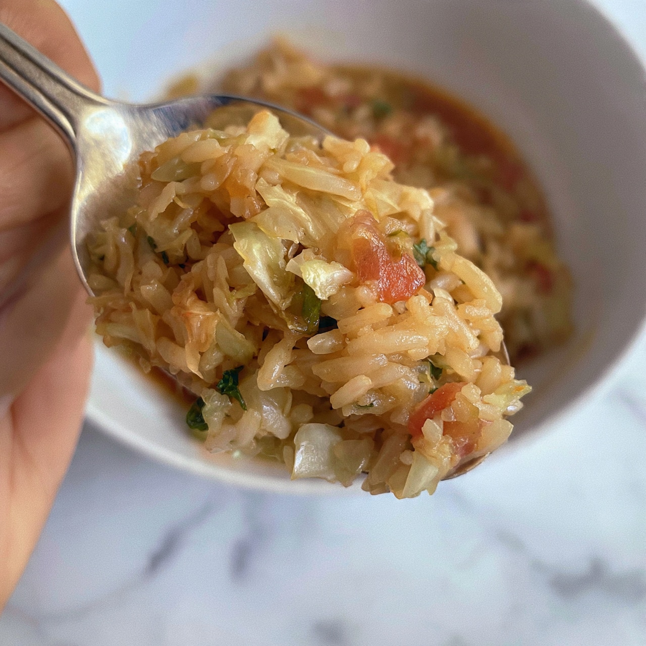 square image The image shows a close-up of a spoon full of cooked rice mixed with small pieces of cabbage, diced tomatoes, and green herbs. The rice is light brown and appears soft, soaking in a reddish-brown sauce. There is a white marbled surface underneath, and the spoon is held by a woman's hand. Photo taken with an iphone --ar 4:5 --v 7