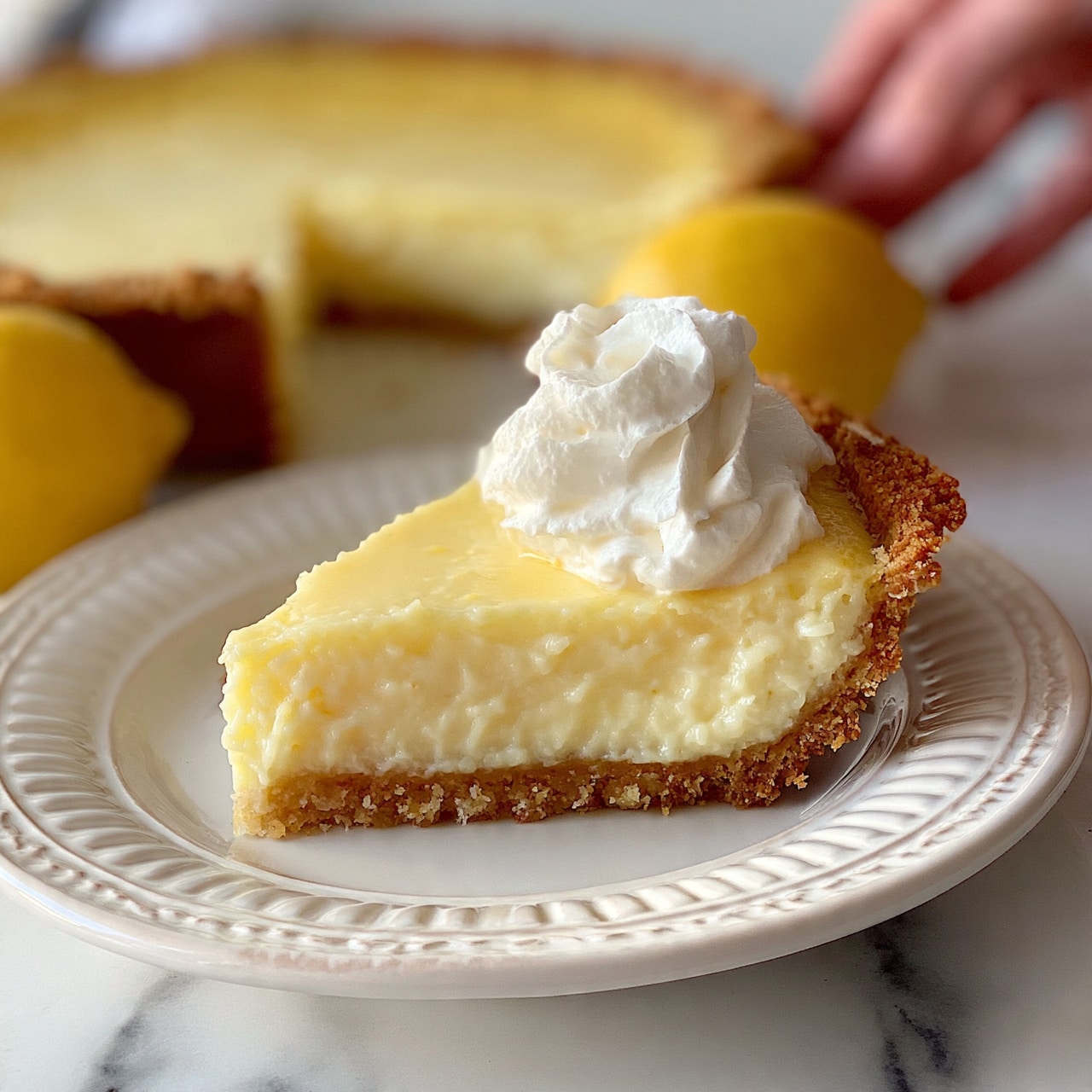 square image A single slice of creamy, pale yellow lemon pie sits on a white ornate ceramic plate with gentle ridges, placed on a white marbled surface. The pie has three visible layers: a crumbly golden brown crust at the bottom, a thick smooth lemon filling in the middle, and a dollop of soft white whipped cream on top. In the background, a whole lemon pie with a smooth, slightly golden top is partially cut and two bright yellow lemons are resting beside it. A woman's hand is reaching towards the pie from the right side. Photo taken with an iphone --ar 4:5 --v 7