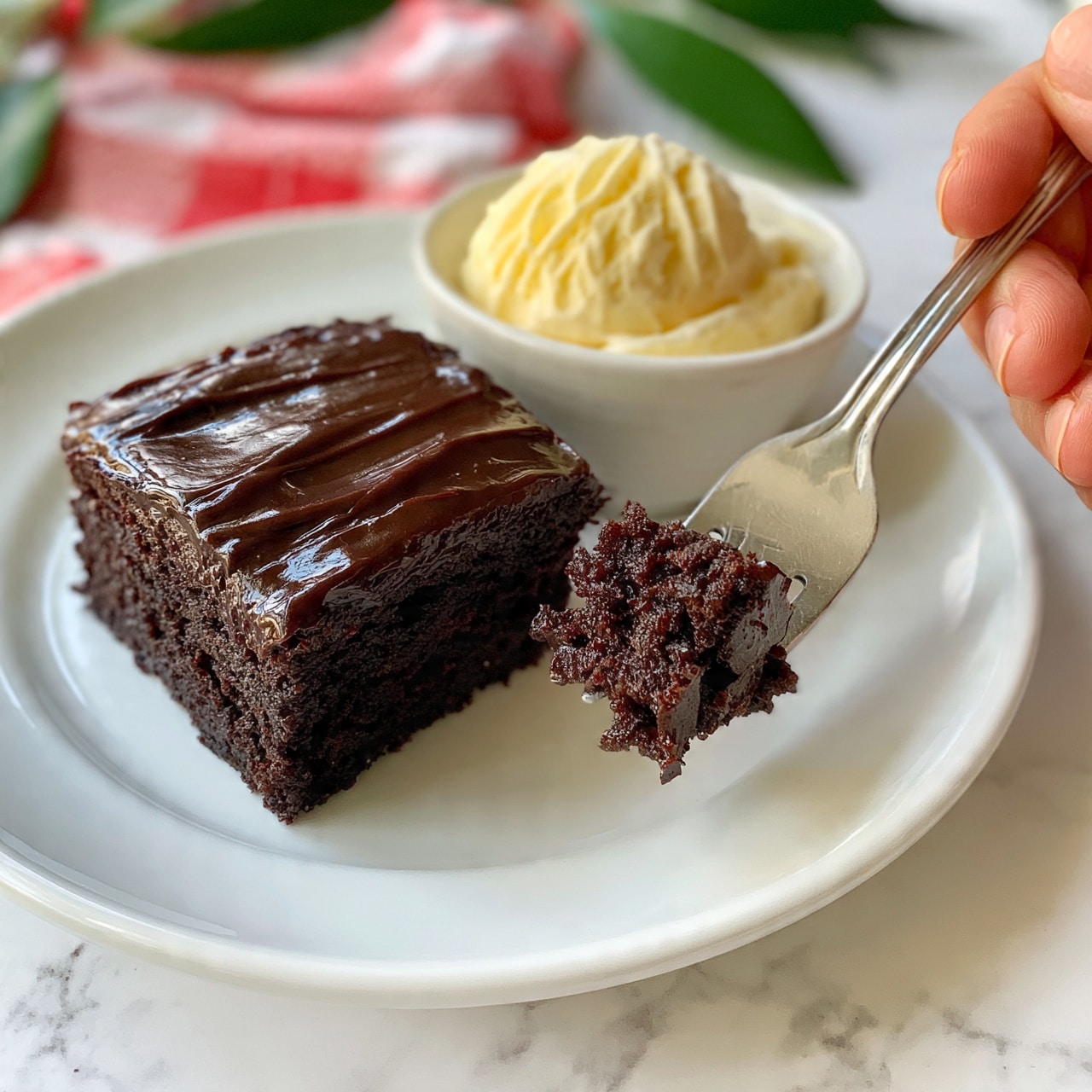 square image A white plate holds a thick, single-layer square piece of dark chocolate cake with a smooth, shiny dark chocolate frosting on top, having slight ridges and a glossy texture. Next to the cake on the plate is a small white bowl filled with a scoop of light yellow vanilla ice cream that looks creamy with some soft ridges. A woman's hand holds a silver fork with a bite-sized piece of the chocolate cake on it, showing the moist and crumbly texture inside. The background shows a blurred red and white checkered cloth and some green leaves, all set on a white marbled surface. photo taken with an iphone --ar 4:5 --v 7