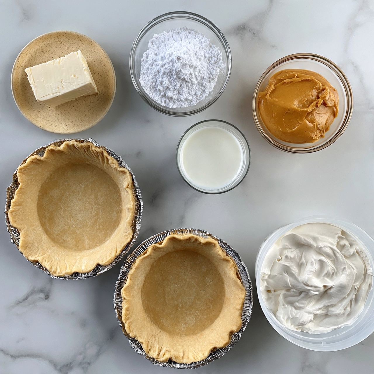 square image The image shows two empty pie crusts placed on a white marbled surface, both with a light tan color and crumbly texture inside shiny silver foil pans. Surrounding the crusts are six small containers with different ingredients: a beige speckled small plate holding a block of off-white cream cheese, a clear glass bowl filled with white powdered sugar, a clear glass bowl filled with smooth, golden-brown peanut butter, a clear glass cup with white milk, and a white plastic container holding thick white whipped cream. The containers and pie crusts are arranged neatly with even spacing. Photo taken with an iphone --ar 4:5 --v 7