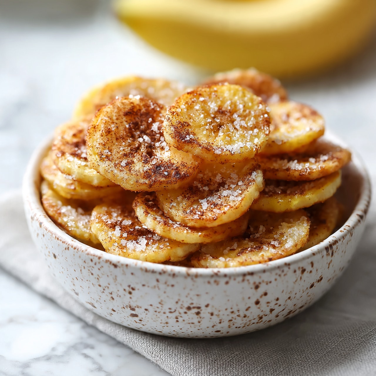 square image A white bowl with brown speckles is filled with many round banana chips stacked in layers. Each chip is golden yellow with a sprinkle of dark brown cinnamon powder and white sugar crystals covering their surfaces. The bowl sits on a light gray cloth on a white marbled surface, and a ripe yellow banana is blurred in the soft background. The warm colors and textures give a crispy and sweet look. Photo taken with an iphone --ar 4:5 --v 7