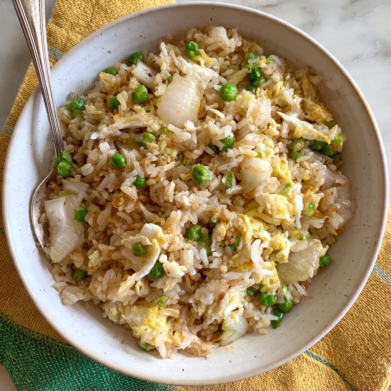 square image A bowl of fried rice with visible layers of light brown cooked rice mixed with small green peas and pale yellow scrambled eggs, along with chunks of soft, translucent white onions. The rice grains are separated and have a slightly oily texture, mixed evenly with the vegetables and eggs. The dish is served in a white bowl with a subtle speckled pattern and contains a silver spoon on the left side inside the bowl. The bowl sits on a yellow and green striped textured cloth over a white marbled surface. Photo taken with an iphone --ar 4:5 --v 7