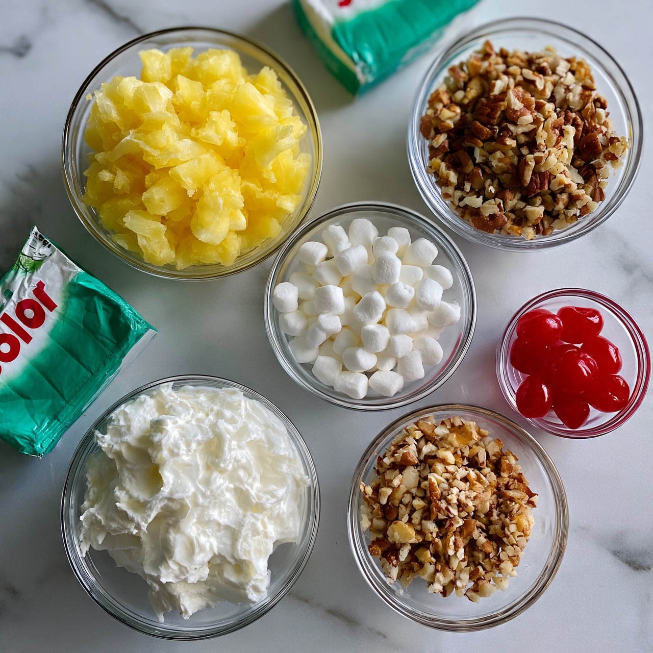 square image The image shows several clear glass bowls and two Jello boxes arranged on a white marbled surface. One bowl is filled with yellow crushed pineapple, another with small white marshmallows, a third with whipped cream, and the fourth contains chopped nuts with a brown and beige color. The two green Jello boxes are placed near the bowls. There's also a small glass bowl with bright red maraschino cherries. The setup is neat and bright, all items are seen from above in a clean, organized way. photo taken with an iphone --ar 4:5 --v 7