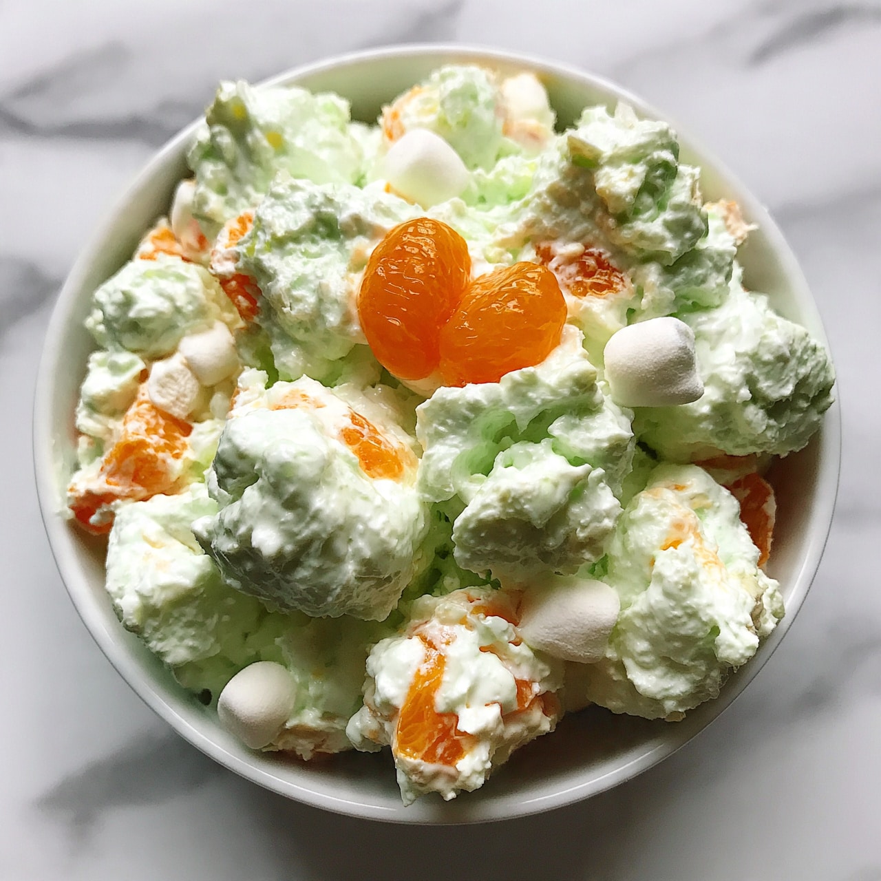 square image The image shows a bowl filled with a creamy, light green salad. The salad has small white marshmallows and bright orange slices mixed inside. The texture looks soft and fluffy, with larger chunks spread throughout. On top of the salad, there are a few small mandarin orange segments placed close together. The bowl is white and sits on a white marbled background. Photo taken with an iphone --ar 4:5 --v 7