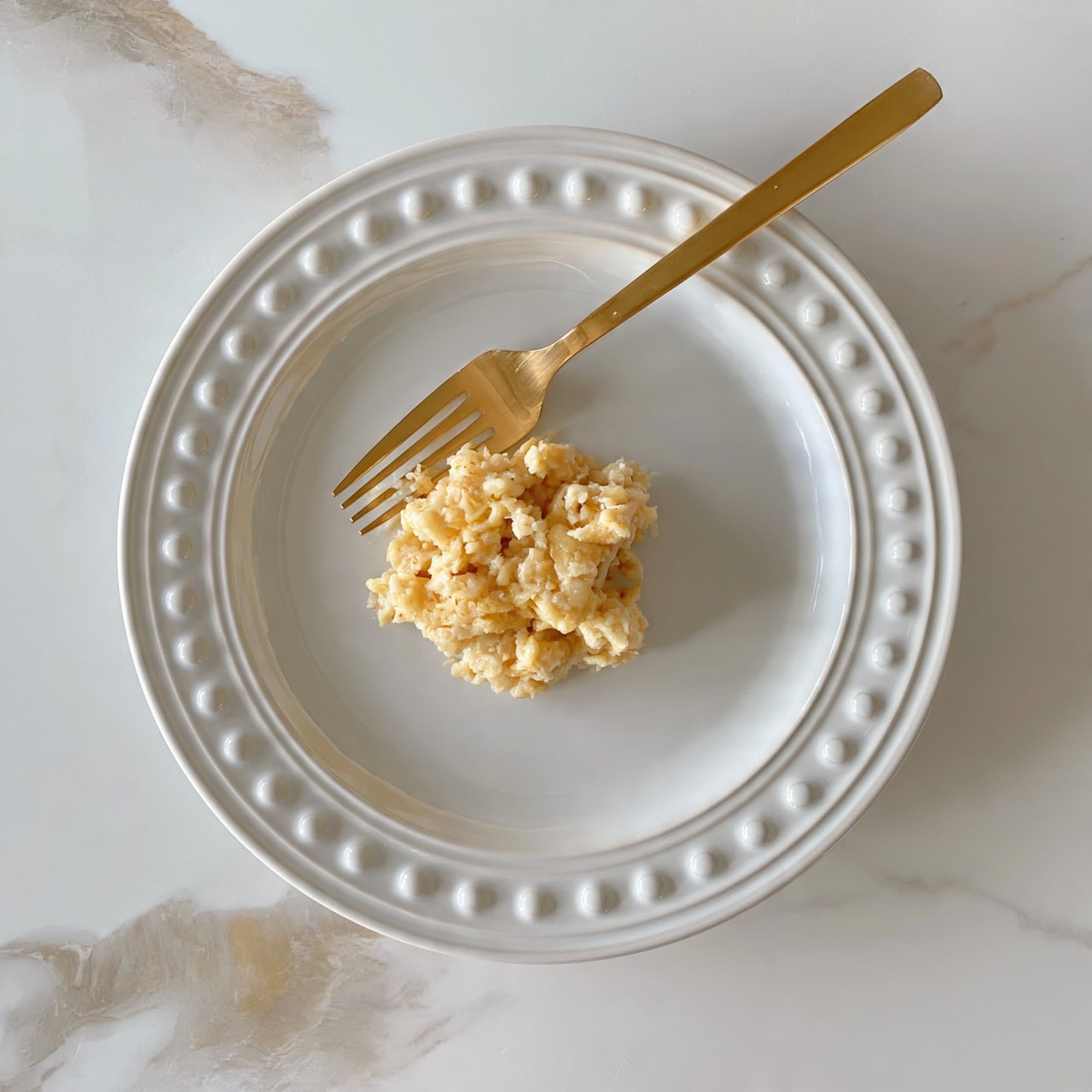 square image The image shows a round white plate with a raised dotted edge. In the middle of the plate is a small mound of mashed bananas with a soft, creamy texture and light golden-yellow color. A gold fork is resting diagonally in the mashed bananas, with its handle pointing toward the top right corner of the image. The plate is placed on a white marbled surface with soft gray and beige tones. The photo taken with an iphone --ar 4:5 --v 7