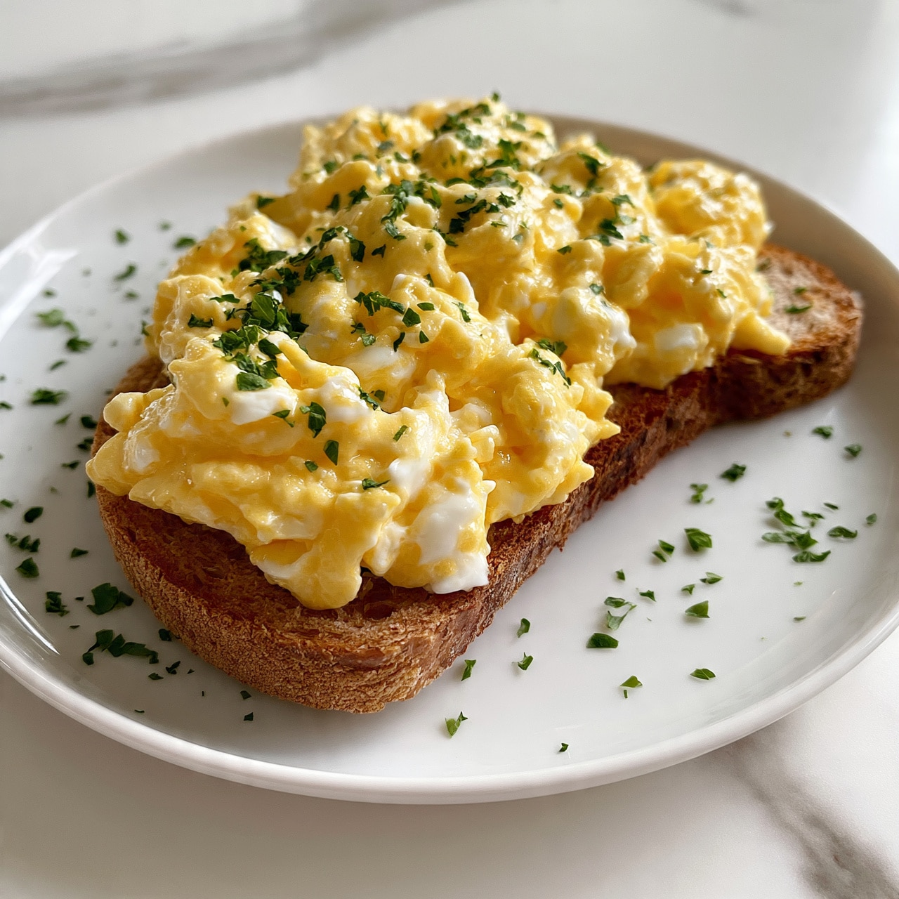 square image The image shows a single slice of toasted brown bread on a white plate with a white marbled surface underneath. On top of the toast is a thick layer of creamy scrambled eggs with a soft, slightly glossy texture and light yellow color mixed with a few white parts. Small chopped green herbs are sprinkled evenly over the eggs and plate edges, adding contrast. The overall look is simple and fresh, with a warm, inviting feel. photo taken with an iphone --ar 4:5 --v 7